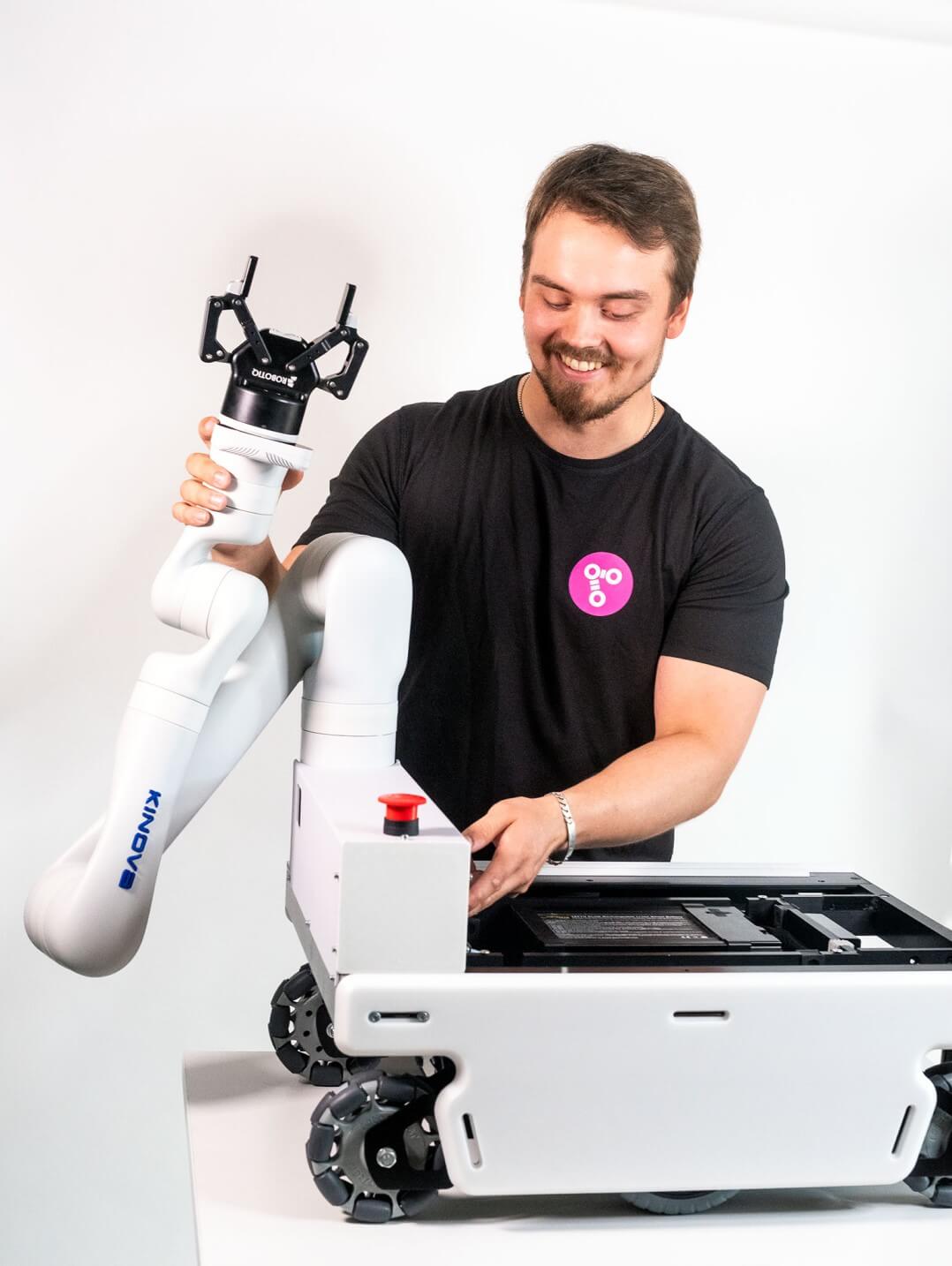 A mobile robot with a white robotic arm and a smiling man standing behind it. The man is wearing a pink Probot logo on his shirt.