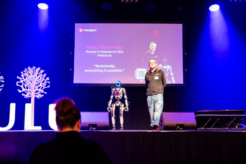 A man and a humanoid robot on stage. In the background, a presentation slide displays the speaker’s name: “Matti Tikanmäki.”