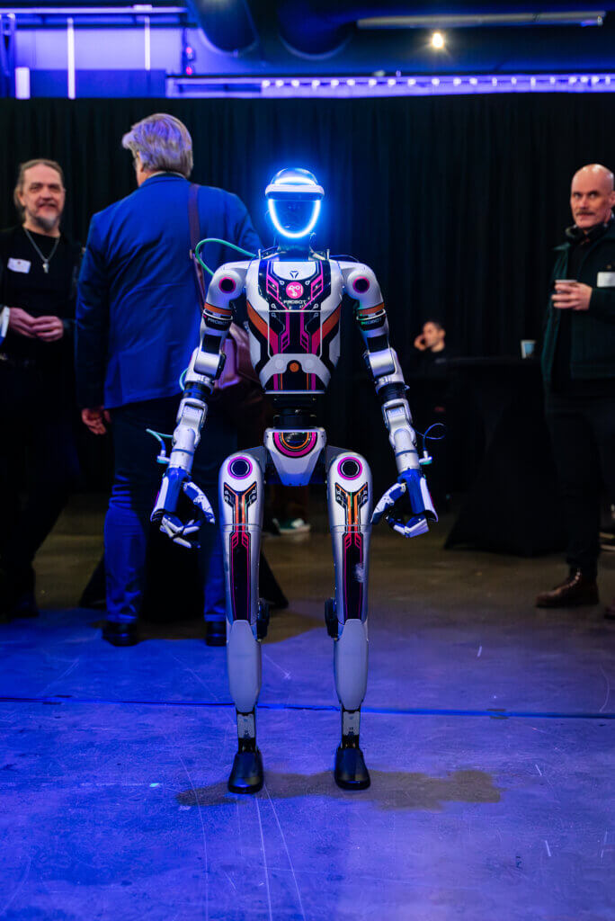 A humanoid robot stands in a dimly lit room, looking directly at the camera. In the background, several men are engaged in conversation.
