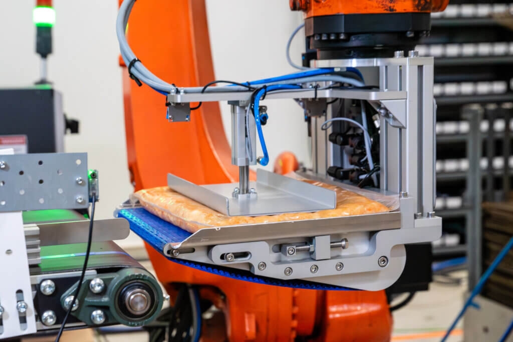 Liukuhihnatarttuja teollisuusrobotin päässä käsittelee vakuumipakattua elintarviketuotetta. Tarttujan sininen kuljetin ja metallirunko näkyvät lähikuvassa.