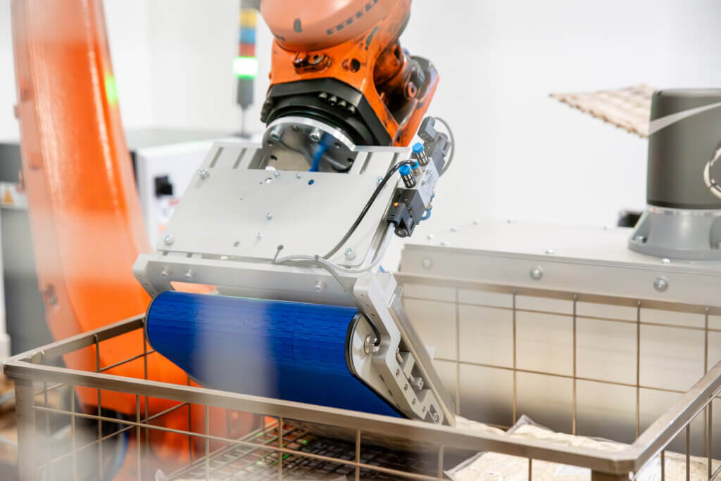 Liukuhihnatarttuja asemoi tuotetta metallisteen autoklaavikoriin. Tarttujan sininen kuljetin ja korin metalliverkkorakenne näkyvät lähikuvassa.