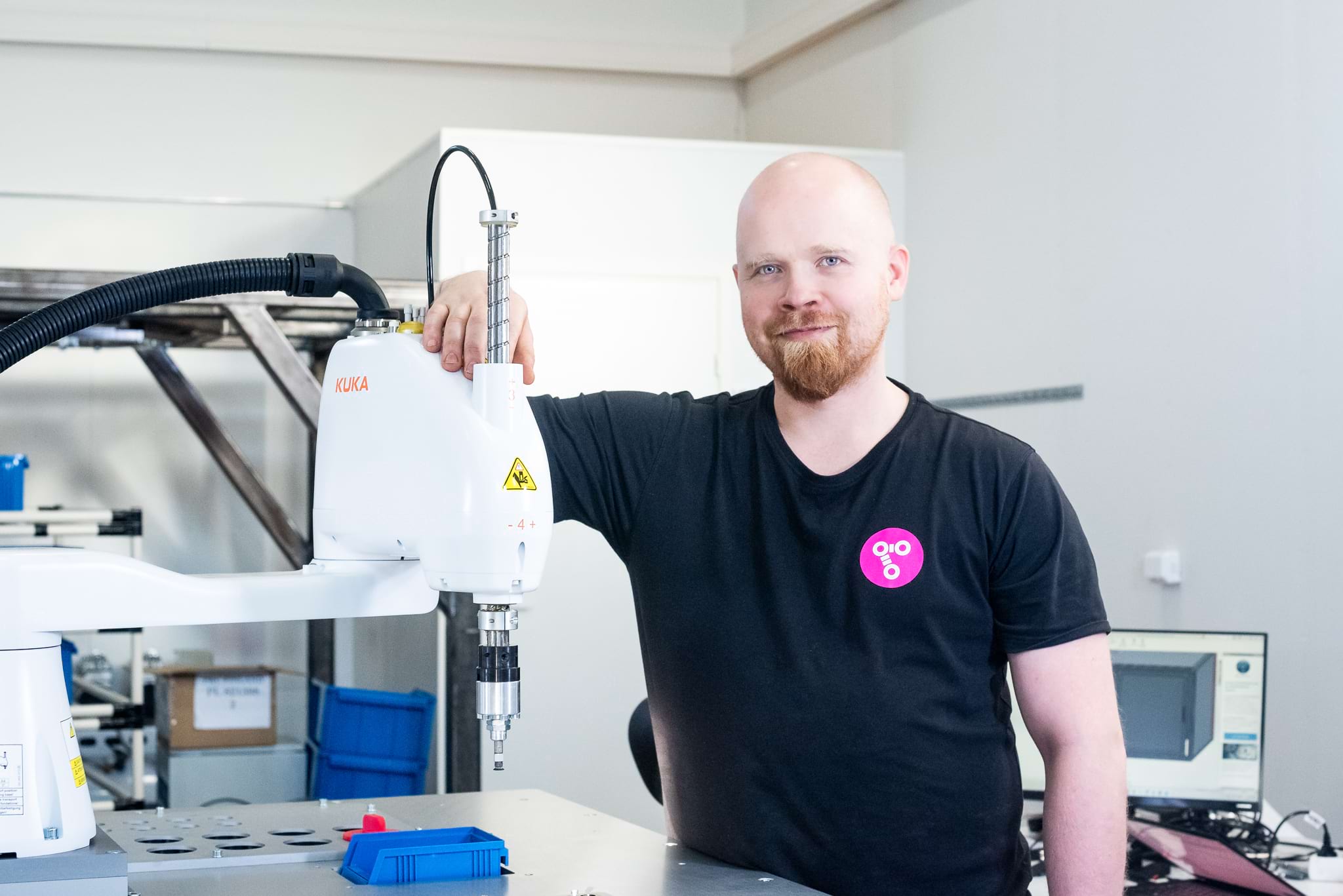 A man dressed in a black t-shirt stands smiling next to a SCARA robot, holding its arm. The robot has 'KUKA' written on it, and in the background, a workspace and a computer are visible.