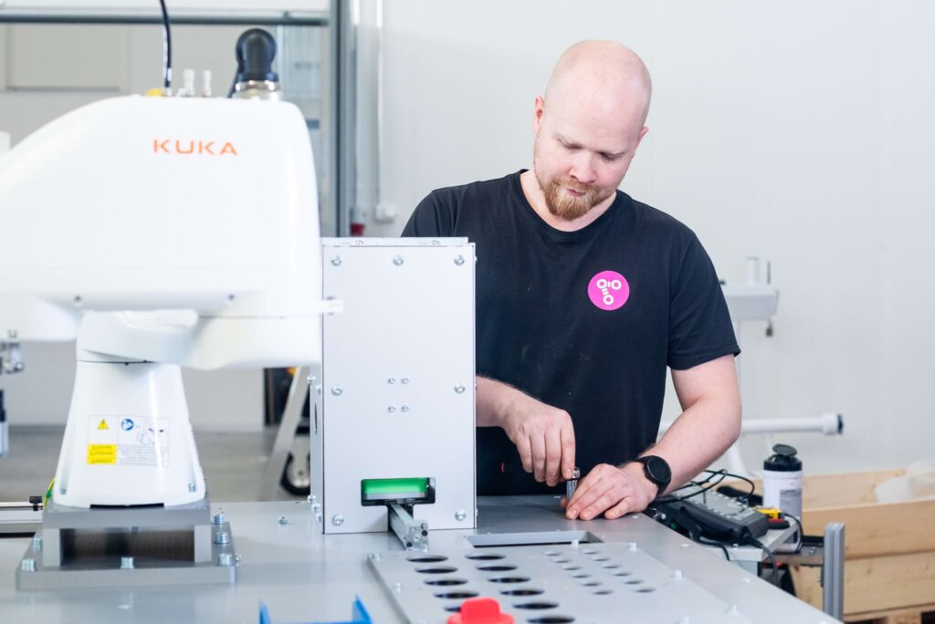 A man wearing a black t-shirt works with a SCARA robot. The robot has the label 'KUKA,' and the man is focused on adjusting a part of the device on a metal workbench.