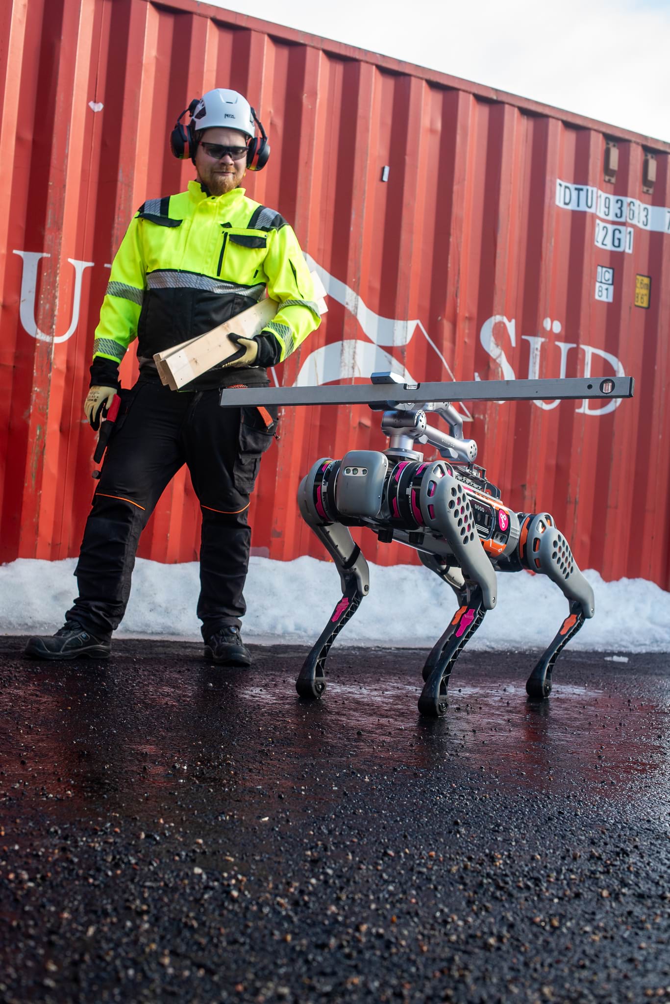 A large robot dog with a robot arm on its back. The robotic arm carries a cotton wool passport. Behind the dog, a construction worker carries a board.