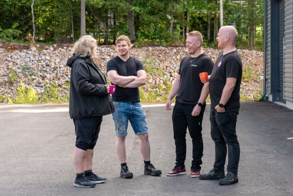 Four men stand outside on an asphalt yard, discussing with a background of forest and rocks. Two of them are holding colorful coffee cups.