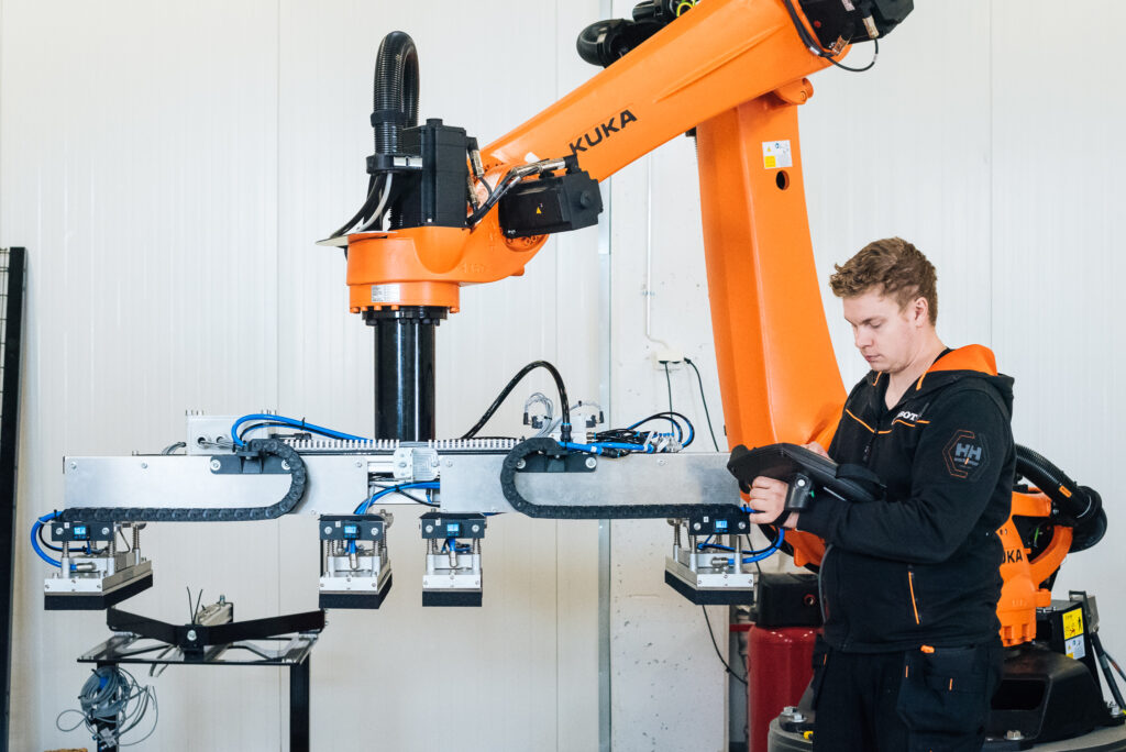 A man is holding a controller in his hands and is thinking about "will robots steal your job?". In the background, an orange KUKA industrial robot.