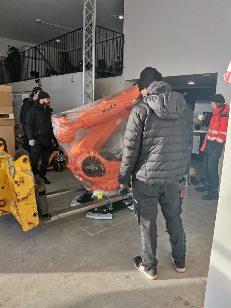 Employees are looking at a wrapped industrial robot, which has arrived at the office.