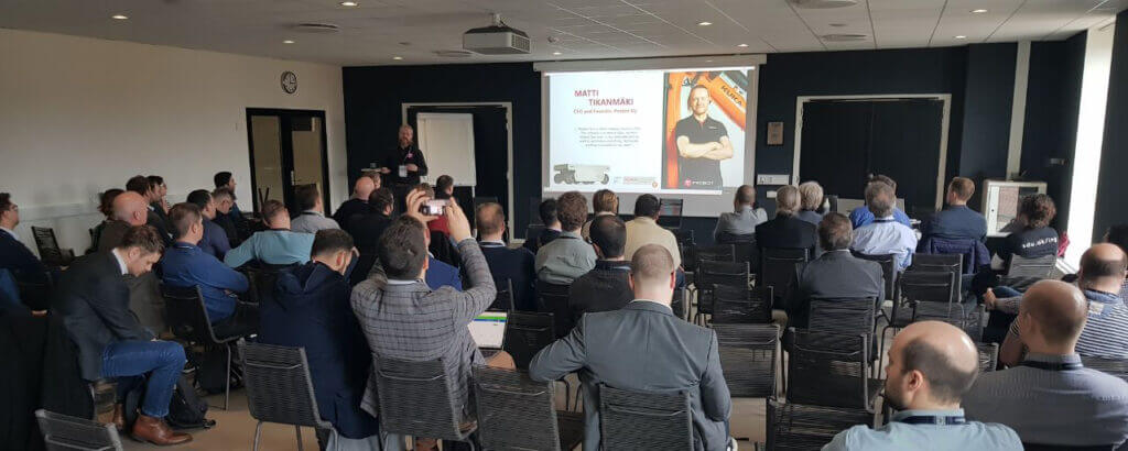 The audience sits with their backs to the camera. In the front part of the space there is a screen with a projected presentation slide. A bearded man stands near the screen. - European Robotics Forum 2023