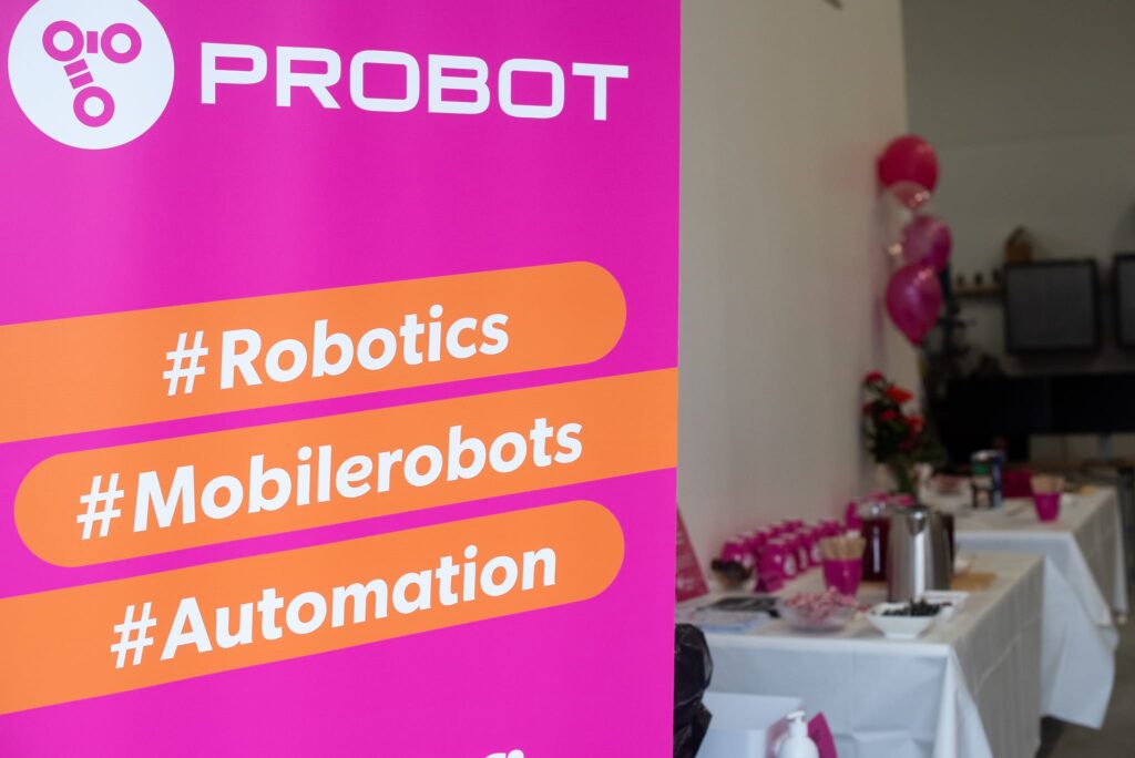 Etualalla pinkki roll up -mainos, jossa robotiikkaa. Taustalla kahvipöytä ja kukkia.