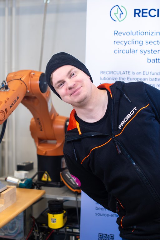 Tiimiläinen hymyilee kameralle robotin edessä. Taustalla EU-projektin roll-up.