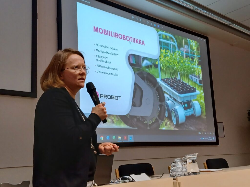 In the foreground, a woman speaking into a microphone at a Forest Seedling Days. In the background is a presentation with information about mobile robotics and a photo of a mobile robot at a forest seedling nursery.