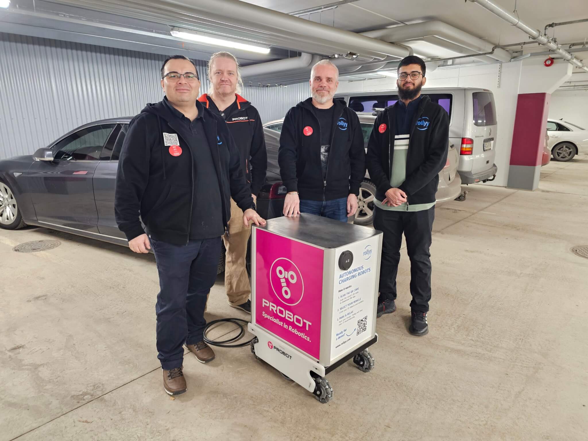 Four members of the Rollyy and Probot team posing together with the autonomous charging robot in a parking garage.