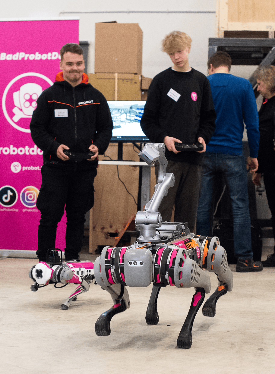 A large and small robot dog run towards the camera. In the background are two men holding robot dog handlers and a pink roll-up with the words #badprobotdog.