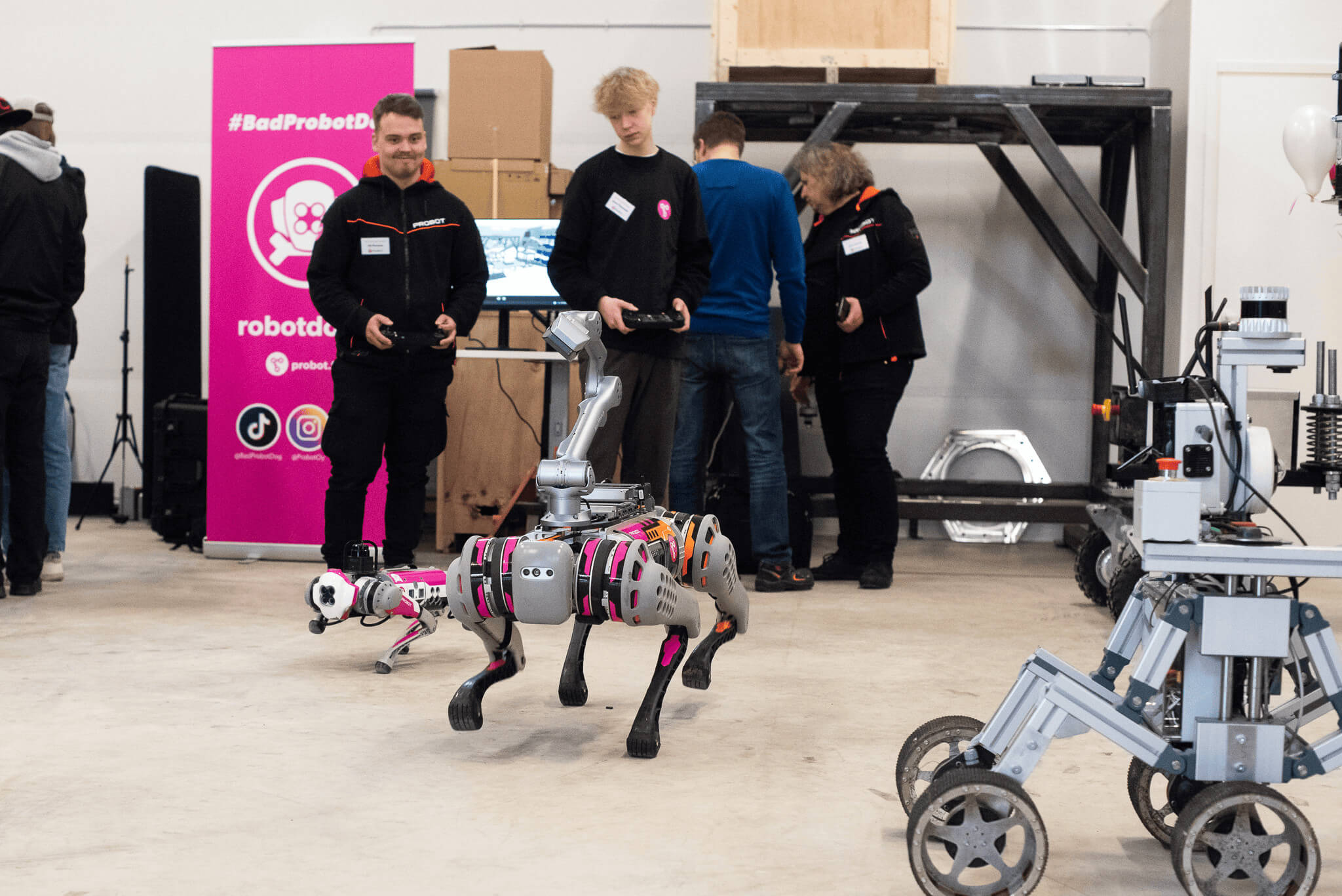 A large and small robot dog run towards the camera. In the background are two men holding robot dog handlers and a pink roll-up with the words #badprobotdog.