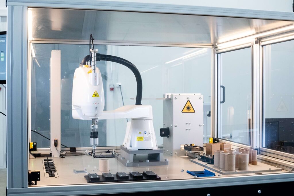 An automation workstation with a white SCARA robot mounted on a metal platform. The table has rows of various parts, such as cardboard tubes and plastic components, along with devices that have warning labels. A glass safety screen and other industrial equipment are visible in the background.