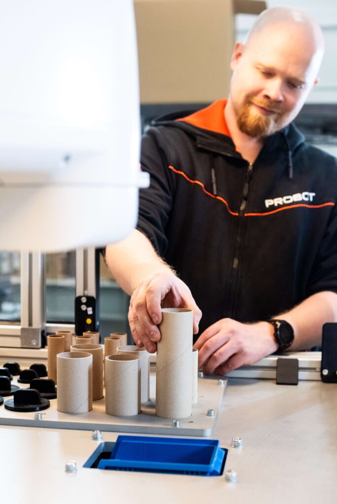 A man wearing a black Probot hoodie works with automation equipment. He is placing cardboard tubes near a robotic device on the table, apparently preparing for a production process.