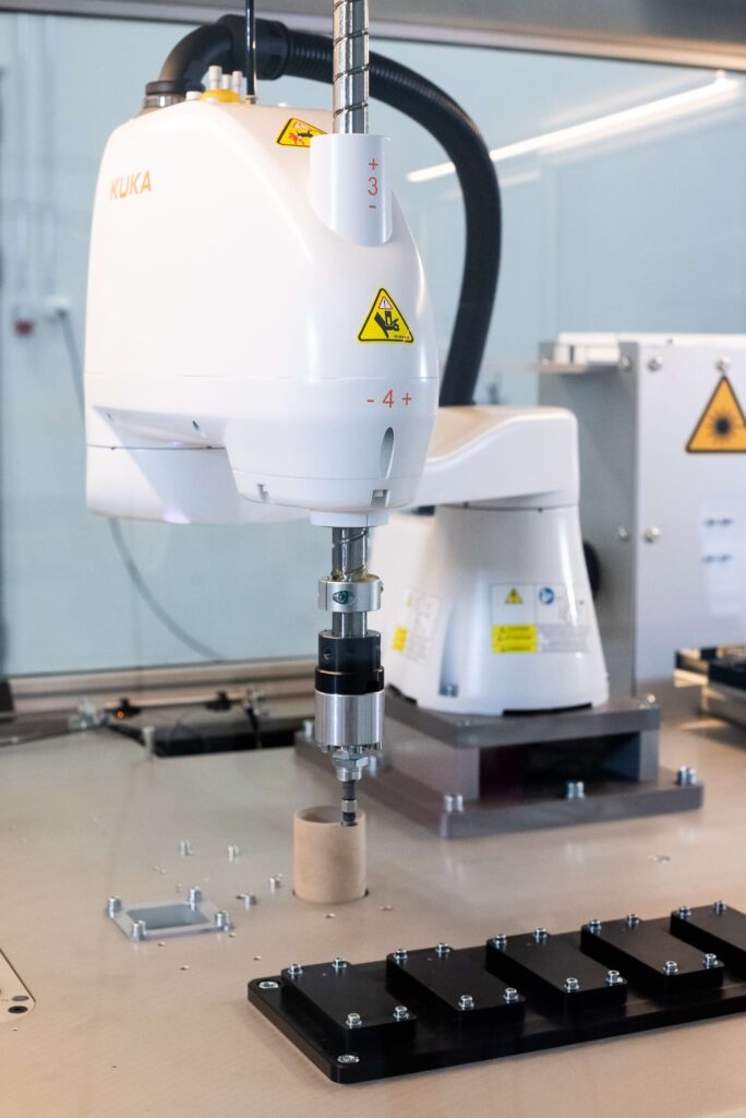 A white KUKA robot working in a glass-enclosed workspace. On the table in front of the robot, there are several components, such as cardboard tubes and metal parts, arranged neatly in order.