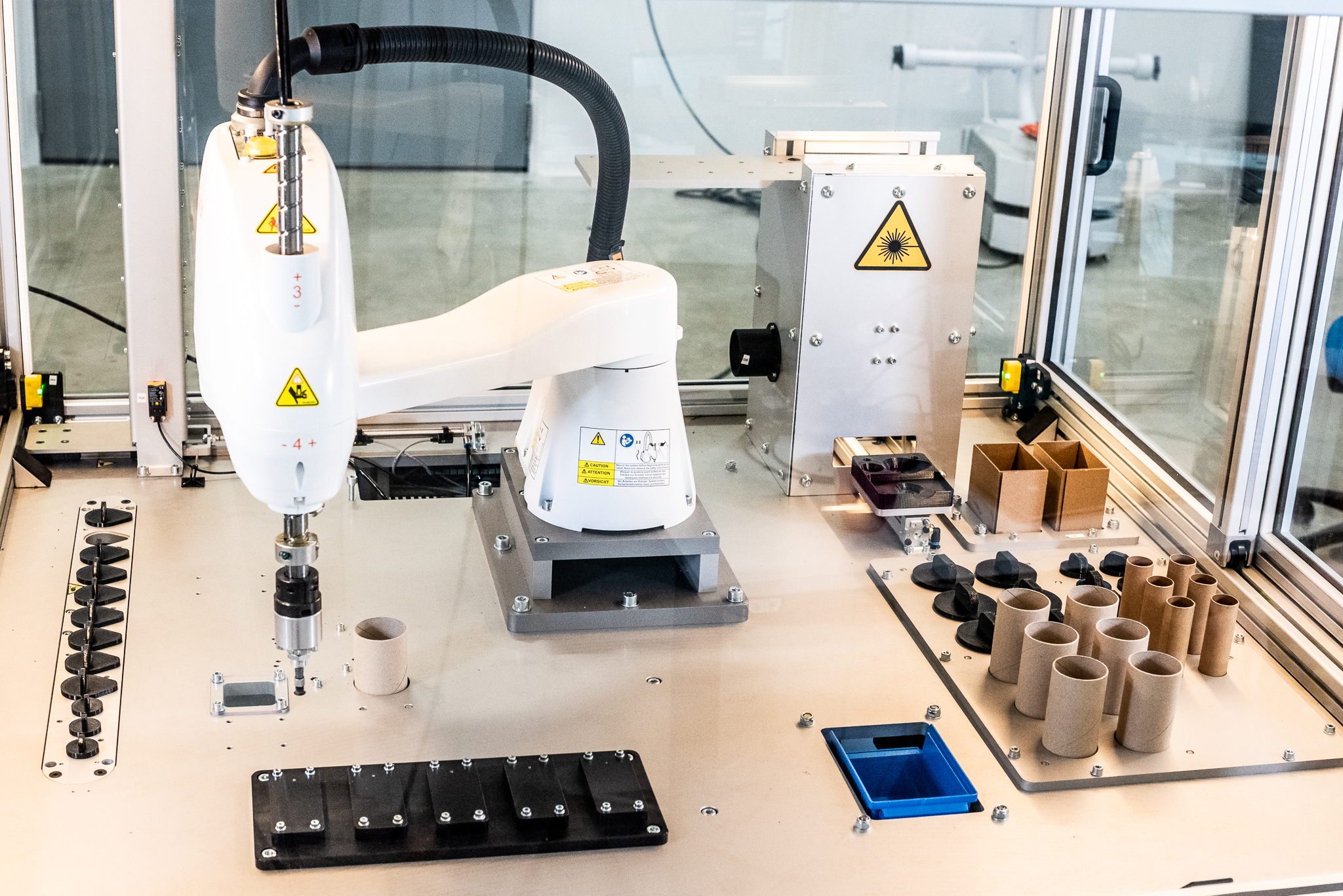 An automation workstation with a white SCARA robot mounted on a metal platform. The table has rows of various parts, such as cardboard tubes and plastic components, along with devices that have warning labels. A glass safety screen and other industrial equipment are visible in the background.