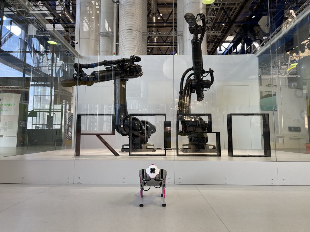 A robot dog stands in front of two large robots. In front of the big robots are movable metal letters that form the word ROBO