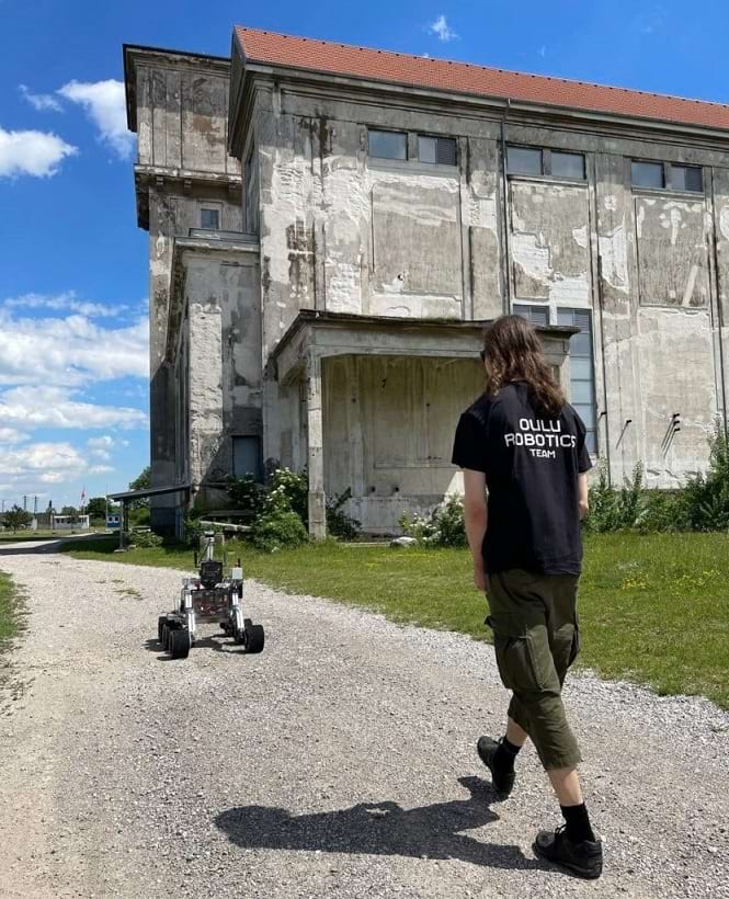 Mies kävelee ulkona robotin takana tätä ohjaten. Hänellä on päällään paita, jossa lukee Oulu Robotics Team.
