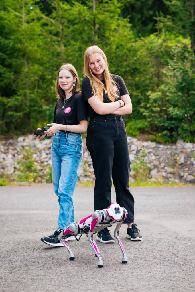 Two smiling girls are standing outside with a robot dog.