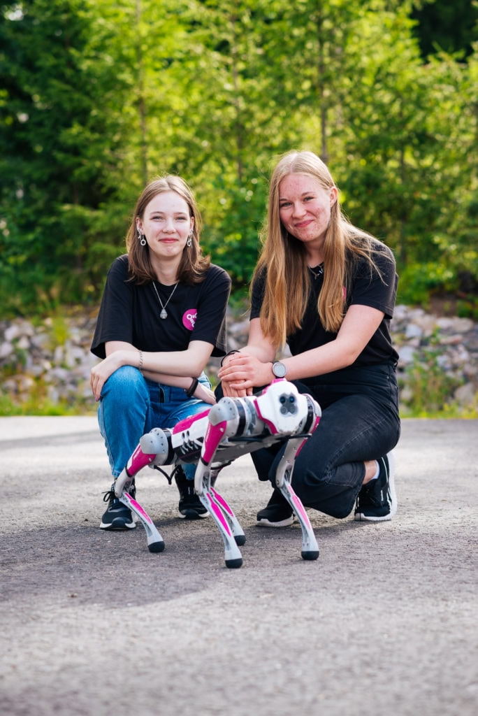 Two girls smile for the camera outdoors together with a robot dog.