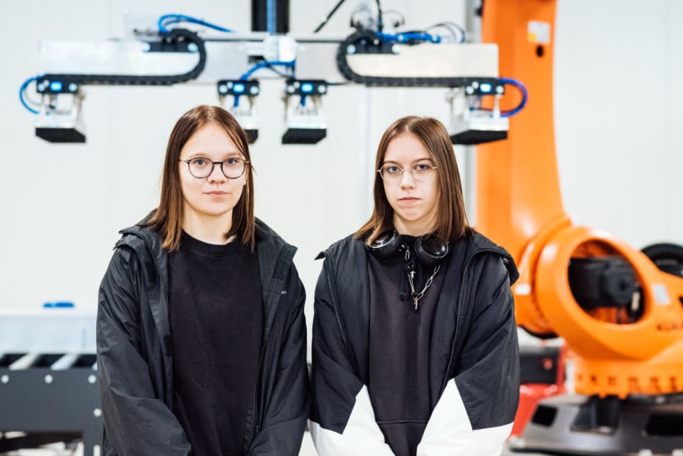 Kaksi nuorta tyttöä seisovat robotin edessä.