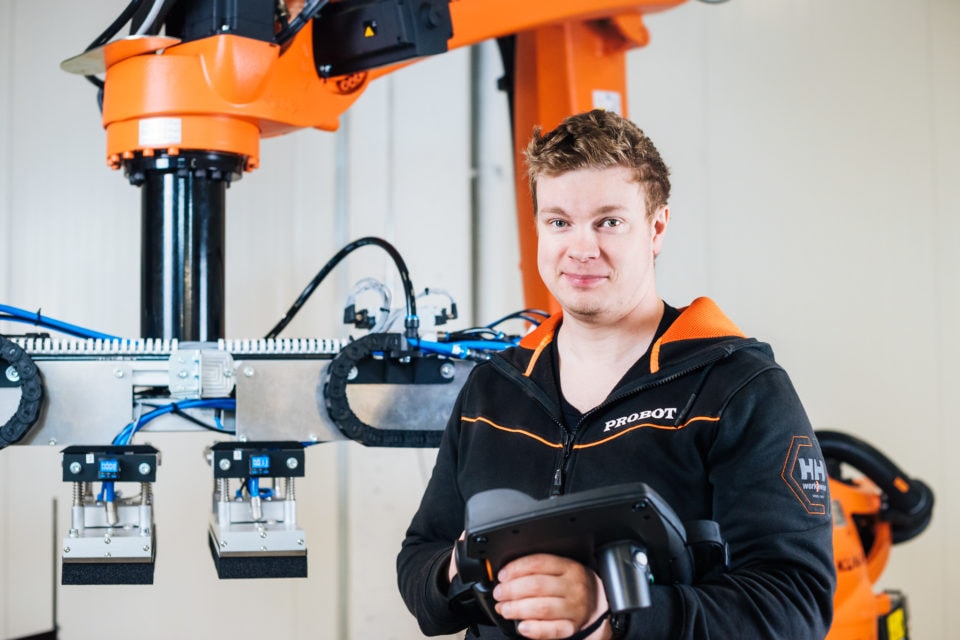 Man stands in front of an industrial robot and smiles at the camera holding a tablet in his hands.