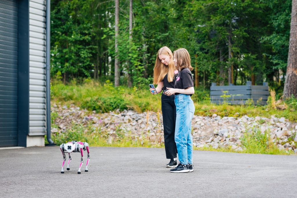 Kaksi nuorta seisovat ulkona robottikoiran kanssa. Toinen nuorista kuvaa robottikoiraa puhelimellaan.