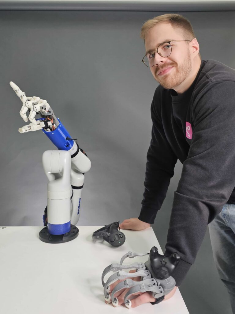 Kinovan käsivarsirobotti Siemensin sormitarttujalla sekä vieressä robotin koodari haptinen hansikas kädessään valmiina näppäryyttä vaativien tehtävien robotisointiin.