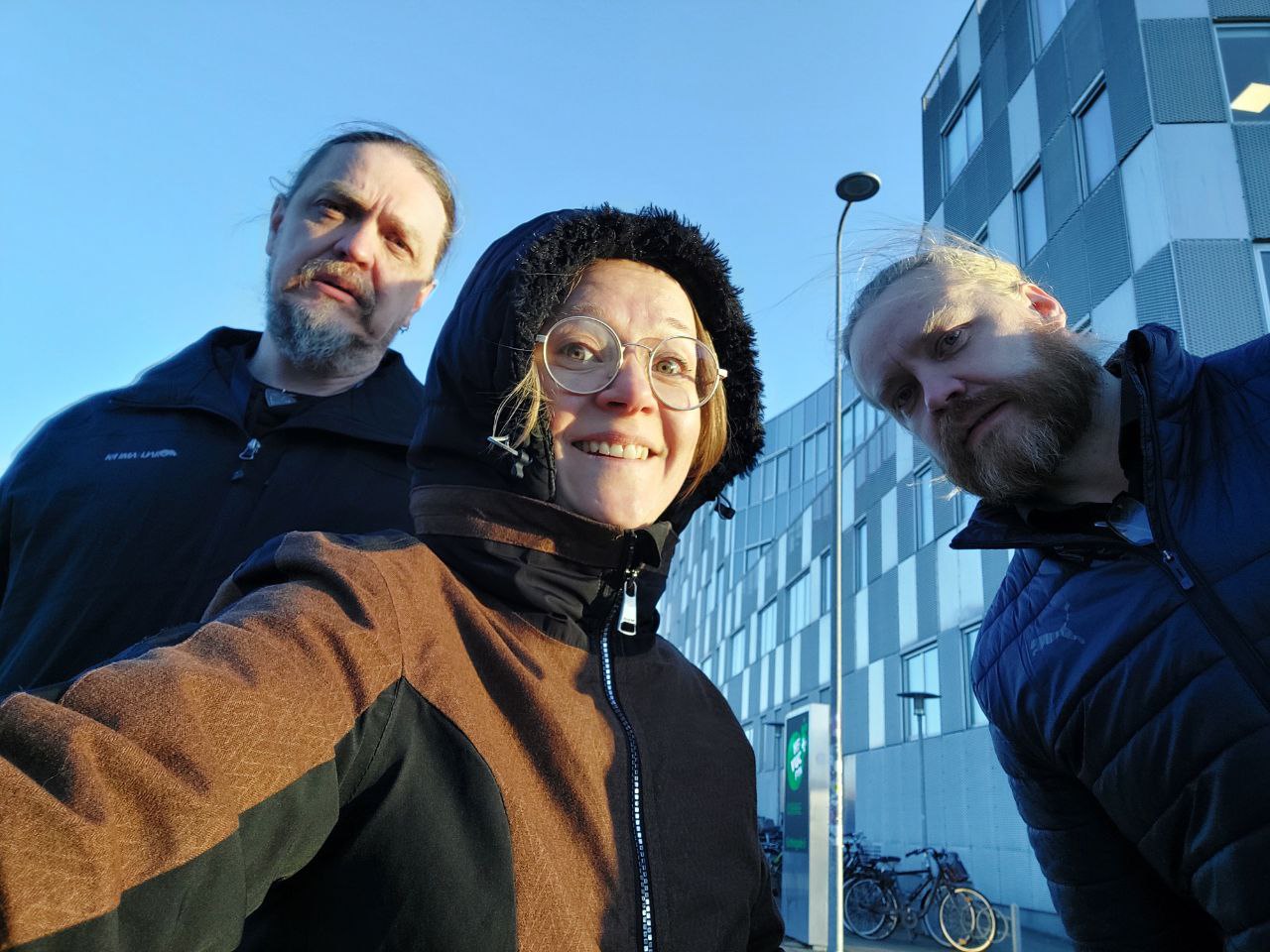 In the close-up, two men and a smiling woman in a hood stand in front of the building. Blue sky in the background.