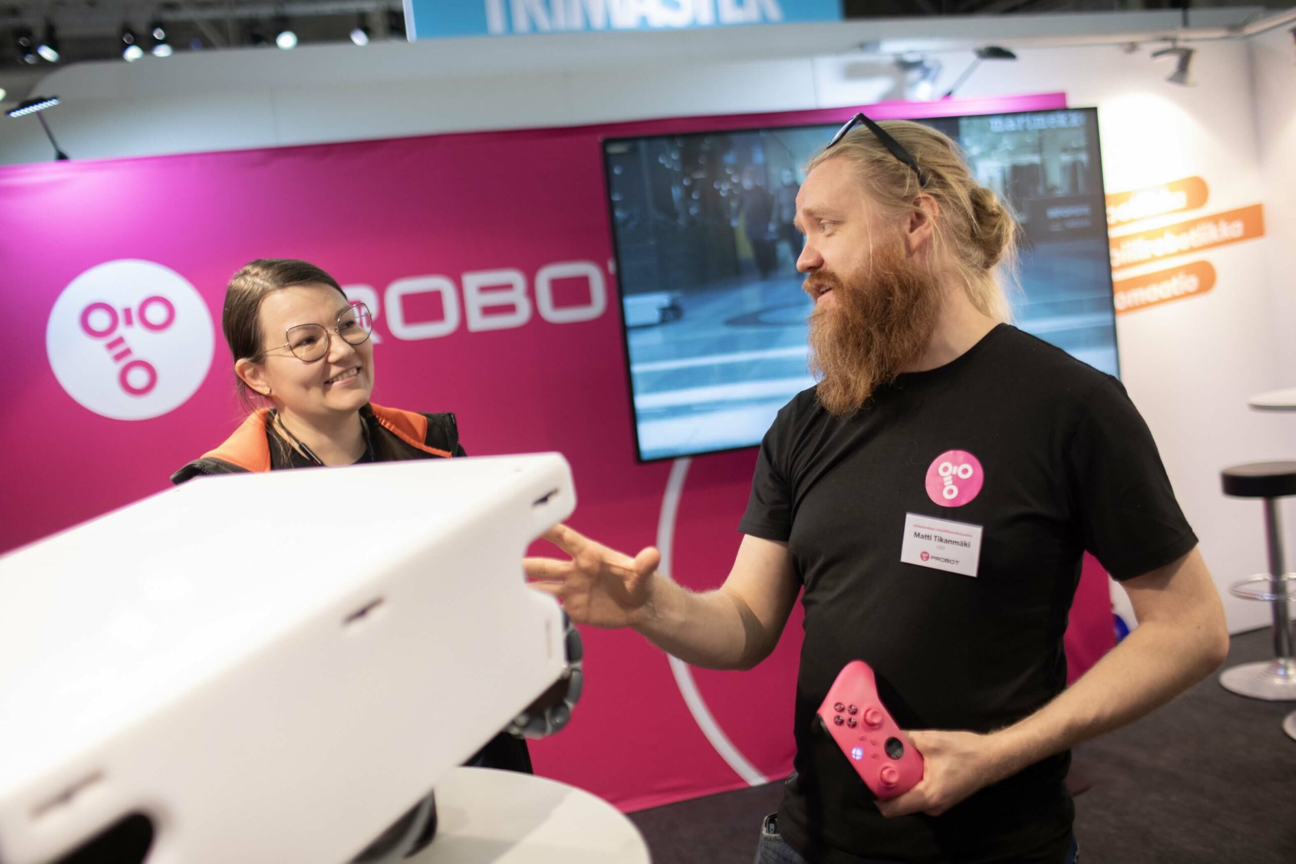 A white mobile robot can be seen in the foreground. Behind it, a man and a woman are talking to each other. In the background, a screen and a pink wall.