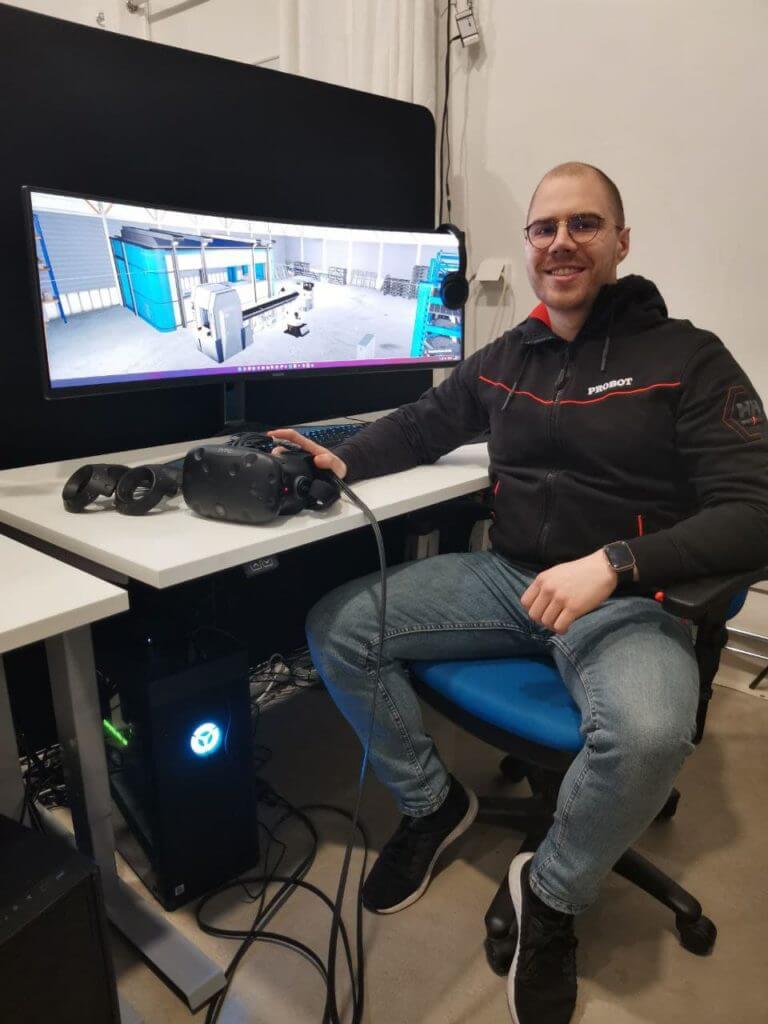 A man sits at his desk and smiles at the camera while holding his hand over virtual reality glasses.