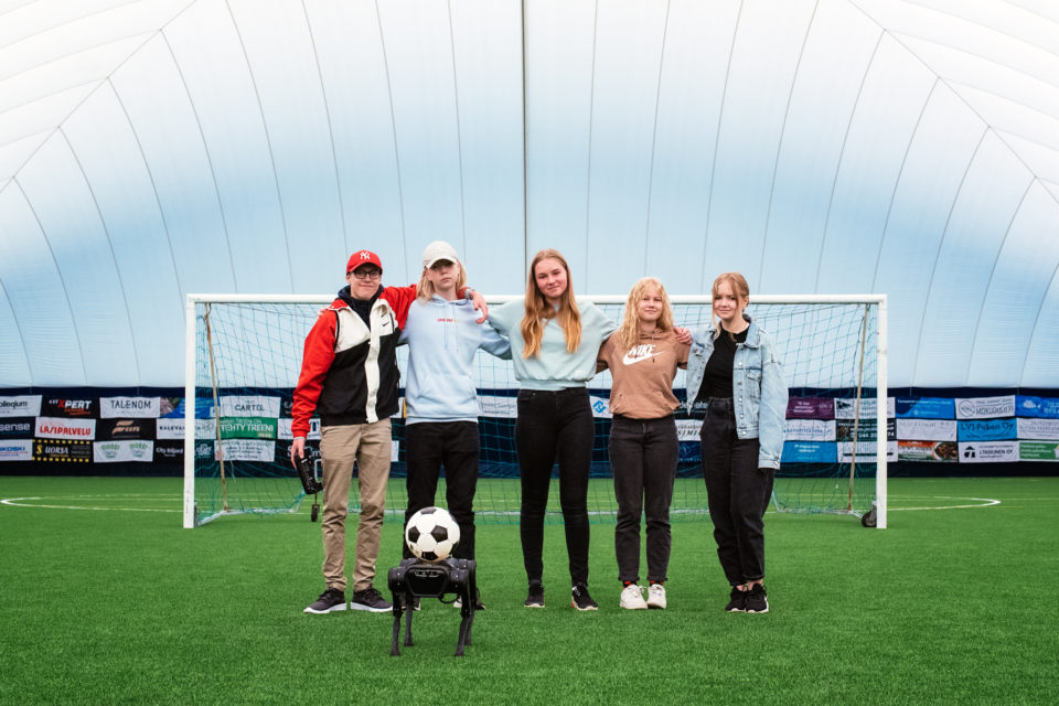 Fife producers of the next generation of robotics standing in line on the fotball field. Thre is a robot dog with a football standing in fron of them.