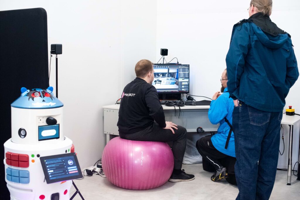 Mies istuu pinkin jumppapallon päällä tietokoneen edessä selkä kameraan. Vieressä toinen mies kyykyssä ja kolmas seisoo. Etualalla robotti.